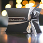 Black gift box with a white ribbon and 'Dukeswickflame' branding on a blurred lights background